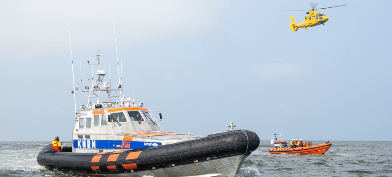 Een grote en kleine reddingboot van de Koninklijke Nederlandse Redding Maatschappij (KNRM) varen op zee naast elkaar. In de lucht vliegt een SAR-helikopter van de Kustwacht.