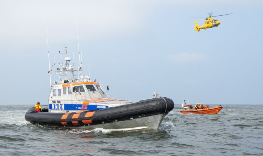 Een grote en kleine reddingboot van de Koninklijke Nederlandse Redding Maatschappij (KNRM) varen op zee naast elkaar. In de lucht vliegt een SAR-helikopter van de Kustwacht.