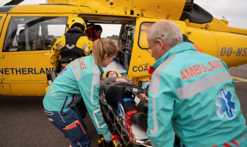 Uit de gele SAR-helikopter wordt voor oefening een brancard met een patiënt gehaald. Deze wordt overgedragen aan ambulancepersoneel.